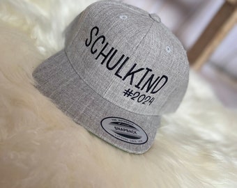 May include: Gray baseball cap with black embroidery that reads "Schulkind #2024". The cap has a black and white label that reads "The Authentic Snapback YUPOONG Since 1974".