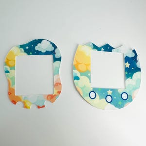 Op de afbeelding: Twee kleurrijke plastic schermbeschermers voor een Tamagotchi-apparaat. De schermbeschermers hebben een blauw, geel en wit wolken- en sterrendesign. Eén schermbeschermer is rond-rechthoekig en de andere is eivormig.