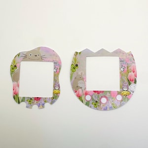 Puede incluir: Dos marcos decorativos con un diseño floral y un personaje de dibujos animados. Los marcos son blancos con un patrón floral rosa, morado y verde. Los marcos tienen una abertura cuadrada blanca en el centro. Los marcos están hechos de papel.