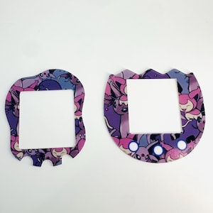 Op de afbeelding: Twee paarse en roze plastic covers voor een draagbare videogameconsole. De covers hebben een vierkante opening in het midden en zijn gevormd als een afgerond rechthoek en een ei.