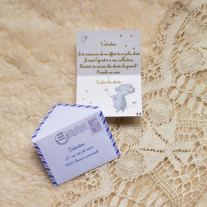 Op de afbeelding: Een witte envelop met een blauwe en paarse rand en een postzegel met de tekst "La F&eacute;e des Dents". De envelop heeft het adres "Valentine, 35 rue du joli dodo, 99600 Beaux sommeils" erop gedrukt. In de envelop zit een gevouwen kaart met een muisillustratie en de tekst "Valentine, Je te remercie de m'offrir ta si jolie dent. Je vais l'ajouter &agrave; ma collection. Bient&ocirc;t, tu auras des dents de grand! Prends-en soin. La f&eacute;e des dents."