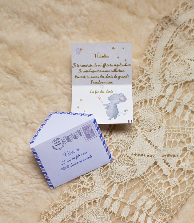 Op de afbeelding: Een witte envelop met een blauwe en paarse rand en een postzegel met de tekst "La F&eacute;e des Dents". De envelop heeft het adres "Valentine, 35 rue du joli dodo, 99600 Beaux sommeils" erop gedrukt. In de envelop zit een gevouwen kaart met een muisillustratie en de tekst "Valentine, Je te remercie de m'offrir ta si jolie dent. Je vais l'ajouter &agrave; ma collection. Bient&ocirc;t, tu auras des dents de grand! Prends-en soin. La f&eacute;e des dents."