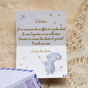Op de afbeelding: Een witte kaart met gouden letters en een kleine illustratie van een muis die een toverstaf vasthoudt met een ster aan het einde. De kaart zegt "Valentine" en "Je te remercie de m'offrir ta si jolie dent. Je vais l'ajouter &agrave; ma collection. Bient&ocirc;t, tu auras des dents de grand! Prends-en soin. La f&eacute;e des dents."