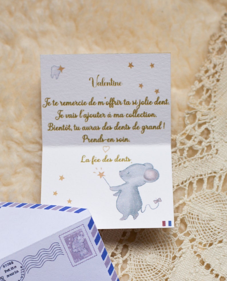 Op de afbeelding: Een witte kaart met gouden letters en een kleine illustratie van een muis die een toverstaf vasthoudt met een ster aan het einde. De kaart zegt "Valentine" en "Je te remercie de m'offrir ta si jolie dent. Je vais l'ajouter &agrave; ma collection. Bient&ocirc;t, tu auras des dents de grand! Prends-en soin. La f&eacute;e des dents."