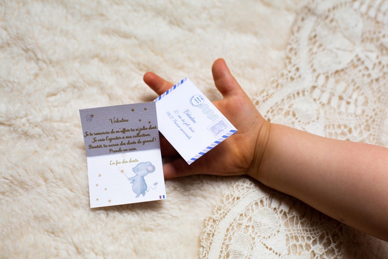Op de afbeelding: Een kleine blauwe envelop met een witte rand en een blauw-witte postzegel. De envelop is geadresseerd aan Valentine en zegt "Aan de tandenfee".  Een kleine kaart met een blauwe muis die een ster vasthoudt en de tekst "La fee des dents" ligt naast de envelop.