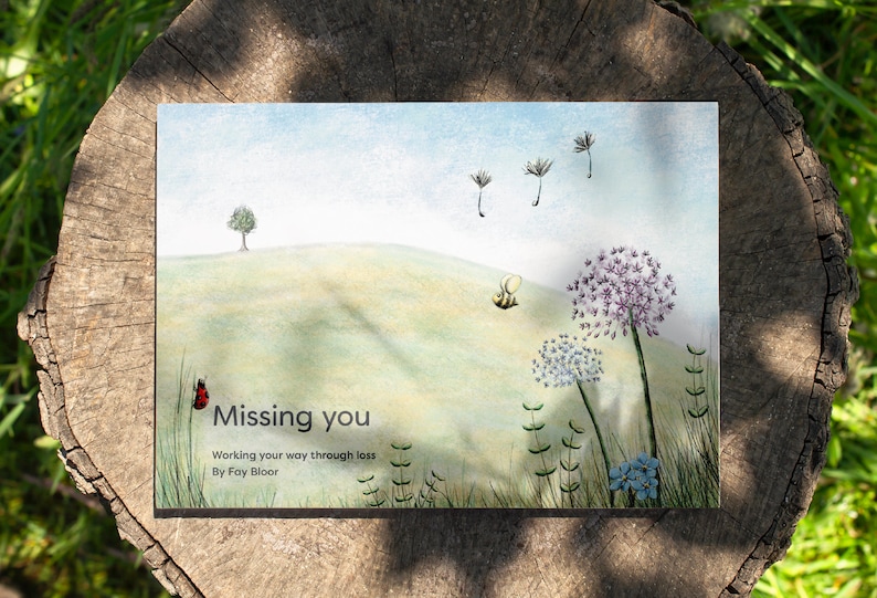 May include: A book cover with a watercolor illustration of a green hill, a small tree, a bee, and dandelions. The title of the book is "Missing you" and the subtitle is "Working your way through loss" by Fay Bloor.