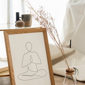 Puede incluir: Una impresi&oacute;n enmarcada con un dibujo lineal minimalista de una persona en una postura de yoga sentada. La obra de arte est&aacute; en un marco de madera clara, exhibida sobre una estera tejida. Una jarra de vidrio con tallos secos y otros objetos decorativos est&aacute;n cerca.