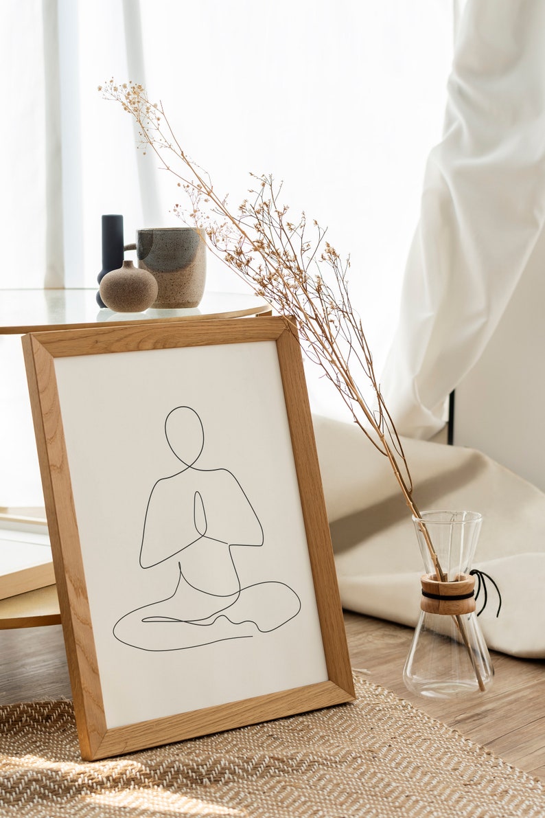 Puede incluir: Una impresi&oacute;n enmarcada con un dibujo lineal minimalista de una persona en una postura de yoga sentada. La obra de arte est&aacute; en un marco de madera clara, exhibida sobre una estera tejida. Una jarra de vidrio con tallos secos y otros objetos decorativos est&aacute;n cerca.