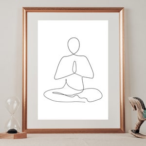 Puede incluir: Una impresi&oacute;n art&iacute;stica minimalista enmarcada. La obra representa a una persona en una postura de yoga sentada con las manos en oraci&oacute;n. La impresi&oacute;n est&aacute; en un marco dorado. Un caballo balanc&iacute;n de madera y un reloj de arena est&aacute;n en el fondo.