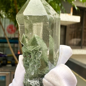 May include: A large, clear quartz crystal with green inclusions. The crystal is faceted and has a pointed tip. It is being held in a white gloved hand.
