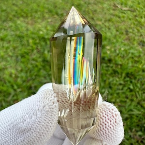 Peut inclure: Une pointe de cristal transparente, facetée, de couleur brun doré avec des reflets arc-en-ciel irisés. Le cristal est tenu dans un gant en tricot blanc.