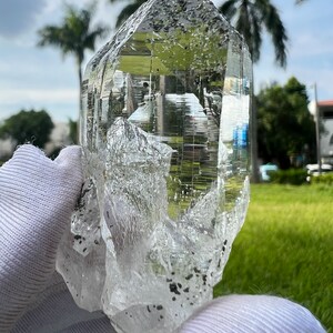 May include: A large, clear quartz crystal with a double termination and inclusions. The crystal is held in a white glove against a green grassy background.