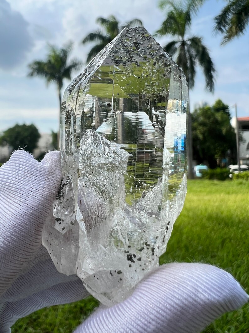 May include: A large, clear quartz crystal with a double termination and inclusions. The crystal is held in a white glove against a green grassy background.
