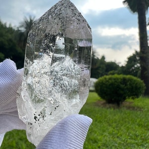 May include: A large, clear quartz crystal with a pointed tip and a rough, textured surface. The crystal is held in a white glove against a blurred background of green grass and trees.