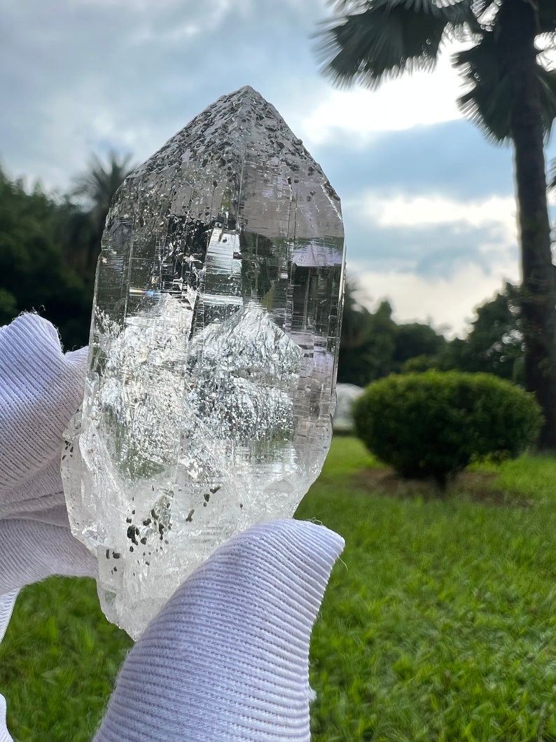 May include: A large, clear quartz crystal with a pointed tip and a rough, textured surface. The crystal is held in a white glove against a blurred background of green grass and trees.
