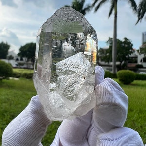 May include: A large, clear quartz crystal with a pointed tip and a flat base. The crystal is held in white gloves against a green grassy background.