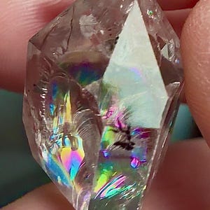 Op de afbeelding: Een helder, gefacetteerd kristalpunt met interne breuken die levendige, veelkleurige regenbooglichtreflecties door de steen creëren. Het transparante mineraal heeft een puntige bovenkant en scherpe randen.