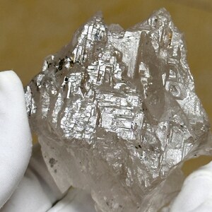 Può includere: Un grande cristallo di diamante chiaro e sfaccettato con una texture ruvida e naturale. Il cristallo è tenuto in una mano guantata.