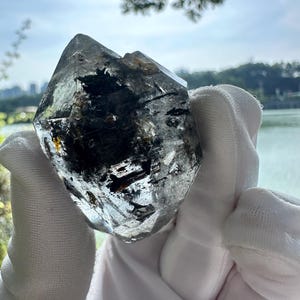 May include: A clear quartz crystal with black inclusions. The crystal is held in a white glove, with a lake and trees in the background. The crystal has a pointed top and a faceted surface.