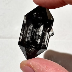Pode incluir: Um cristal de quartzo preto de terminação dupla com forma prismática hexagonal. O cristal apresenta um corpo escuro e opaco com bordas translúcidas e uma pequena protuberância cristalina secundária em um dos seus lados.