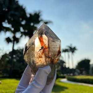 May include: A large, clear quartz crystal with reddish-brown inclusions. The crystal is shaped like a point and is being held in a white gloved hand.