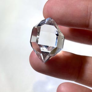 May include: A clear, faceted crystal with a symmetrical, double-terminated shape. The crystal is held between fingers, showcasing its transparency and geometric design. The crystal's facets reflect light, creating a sparkling effect.