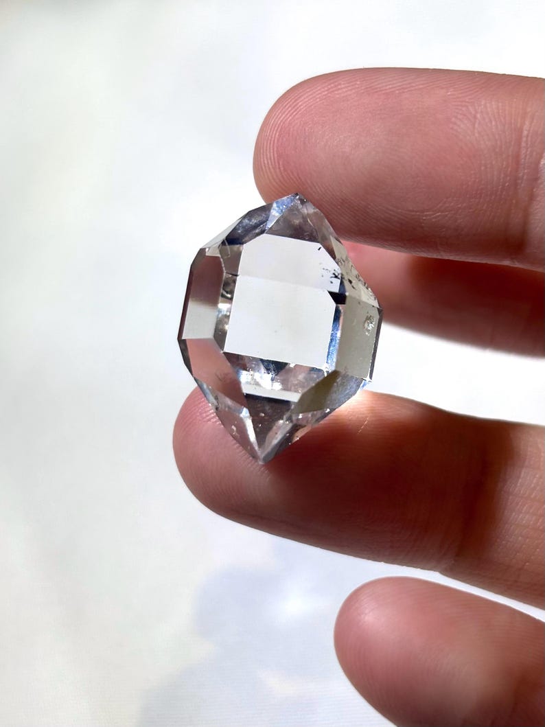 May include: A clear, faceted crystal with a symmetrical, double-terminated shape. The crystal is held between fingers, showcasing its transparency and geometric design. The crystal's facets reflect light, creating a sparkling effect.