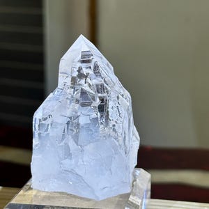 May include: A clear quartz crystal with a pointed top and a rough, textured surface. The crystal is mounted on a clear acrylic base, showcasing its natural beauty. The crystal's facets reflect light, creating a sparkling effect. This is a natural gemstone.