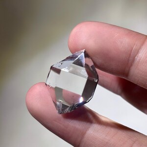 May include: A clear, faceted crystal held between fingers. The crystal has a geometric shape with multiple facets, reflecting light. The overall appearance is transparent with a slight gray hue, suggesting it is a quartz crystal.