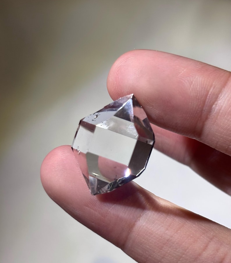 May include: A clear, faceted crystal held between fingers. The crystal has a geometric shape with multiple facets, reflecting light. The overall appearance is transparent with a slight gray hue, suggesting it is a quartz crystal.