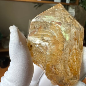 May include: A large, clear quartz crystal with a golden hue and internal inclusions. The crystal is faceted and has a pointed tip. It is being held in a white gloved hand.
