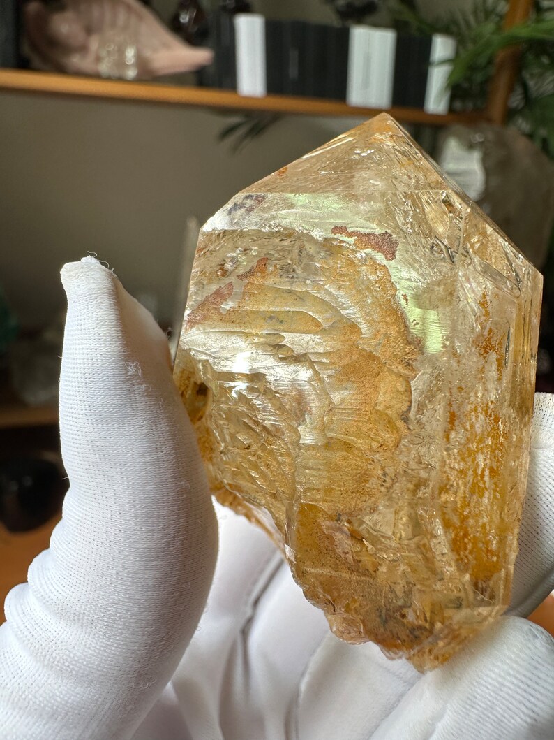 May include: A large, clear quartz crystal with a golden hue and internal inclusions. The crystal is faceted and has a pointed tip. It is being held in a white gloved hand.