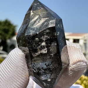 May include: A large, clear quartz crystal with black inclusions. The crystal is double-terminated and has a pointed shape.