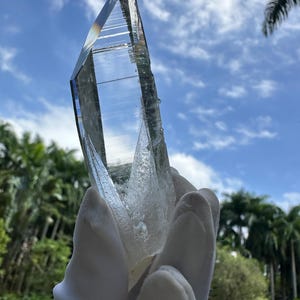 Pode incluir: Um grande cristal de quartzo transparente, segurado contra um céu azul com nuvens brancas e árvores verdes exuberantes. O cristal é facetado e reflete a luz, mostrando sua transparência e beleza natural.
