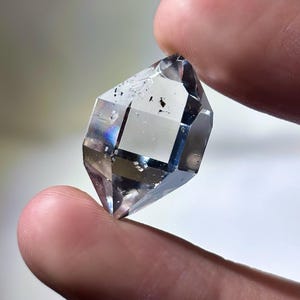 May include: A clear, faceted crystal with a geometric shape. The crystal is held between two fingers, showcasing its transparency and internal structure. The crystal has a few dark inclusions and displays a subtle rainbow iridescence.