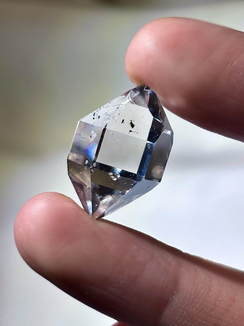 May include: A clear, faceted crystal with a geometric shape. The crystal is held between two fingers, showcasing its transparency and internal structure. The crystal has a few dark inclusions and displays a subtle rainbow iridescence.