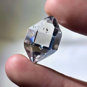 May include: A clear, faceted crystal with a geometric shape. The crystal is held between two fingers, showcasing its transparency and internal structure. The crystal has a few dark inclusions.