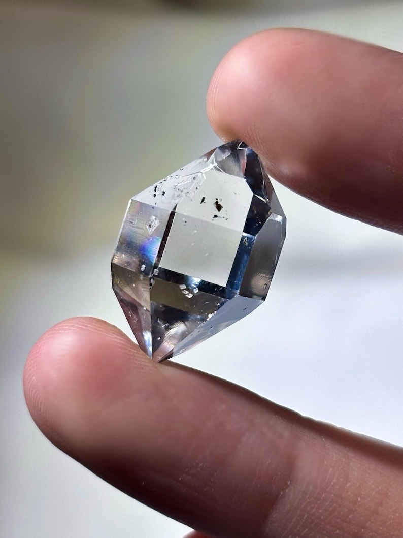 May include: A clear, faceted crystal with a geometric shape. The crystal is held between two fingers, showcasing its transparency and internal structure. The crystal has a few dark inclusions.