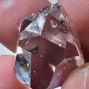 May include: A clear, faceted crystal with multiple geometric faces. The crystal is held between fingers, showcasing its transparent quality and internal inclusions. The light refracts through the crystal, creating highlights and shadows, emphasizing its sharp edges and overall shape.