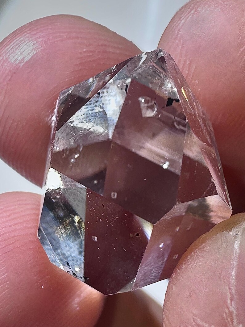 May include: A clear, faceted crystal with multiple geometric faces. The crystal is held between fingers, showcasing its transparent quality and internal inclusions. The light refracts through the crystal, creating highlights and shadows, emphasizing its sharp edges and overall shape.