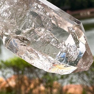 Op de afbeelding: Een heldere kwartskristal met een veelzijdig, getextureerd oppervlak. Het kristal heeft een natuurlijke, ruwe uitstraling met een lichtbruine tint en een puntige punt. De achtergrond is wazig, met groen gebladerte en water zichtbaar.
