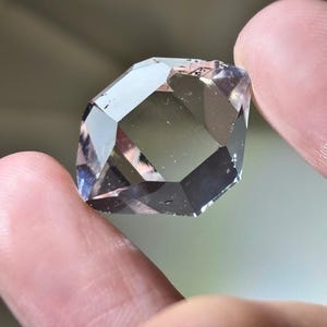 May include: A faceted, clear gemstone with a geometric shape. The gem is held between two fingers, showcasing its multiple reflective surfaces. The stone has a pale, smoky color with hints of purple and gray, catching the light.