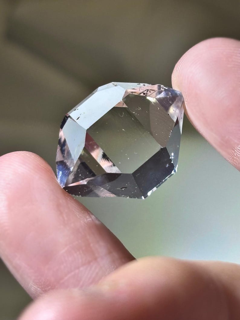 May include: A faceted, clear gemstone with a geometric shape. The gem is held between two fingers, showcasing its multiple reflective surfaces. The stone has a pale, smoky color with hints of purple and gray, catching the light.