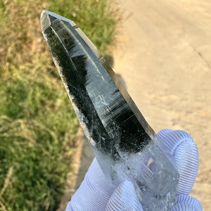 May include: A clear quartz crystal with dark green inclusions. The crystal is held in a white gloved hand. The crystal is a long, pointed shape, with a faceted tip. The crystal is partially transparent.