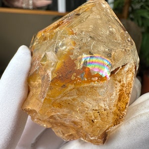 May include: A large, clear quartz crystal with a golden hue and iridescent rainbow inclusions. The crystal has a rough, natural surface and is a beautiful example of a raw gemstone.