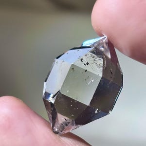 May include: A faceted, clear crystal with a smoky hue is held between two fingers. The crystal's surface reflects light, showcasing its geometric cuts and internal inclusions. The overall color is a mix of clear and smoky gray.