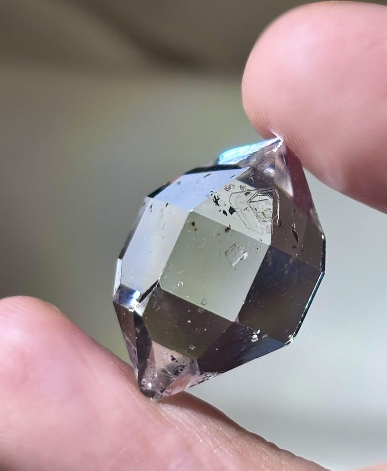 May include: A faceted, clear crystal with a smoky hue is held between two fingers. The crystal's surface reflects light, showcasing its geometric cuts and internal inclusions. The overall color is a mix of clear and smoky gray.