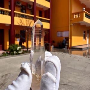 May include: A clear quartz crystal point, held in a gloved hand, with a natural, rough base and a sharply defined tip. The crystal is transparent, allowing light to pass through, and is set against a blurred outdoor background.