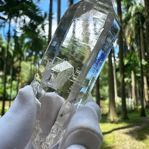 Könnte beinhalten: Ein großer, klarer Quarz-Kristall mit einer Spitze und einer kleineren Kristallformation. Der Kristall wird in einer behandschuhten Hand vor dem Hintergrund grüner Bäume und blauem Himmel gehalten.