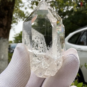 Op de afbeelding: Een heldere bergkristal punt met een facetvormige, geometrische vorm. Het kristal wordt vastgehouden in een witte handschoen, wat de transparantie en natuurlijke textuur benadrukt. De facetten van het kristal vangen het licht op en creëren een sprankelend effect.